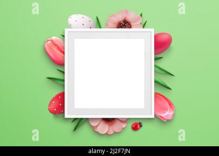 Blank picture frame surrounded by Easter eggs and flowers on green surface. Top view, flat lay composition Stock Photo