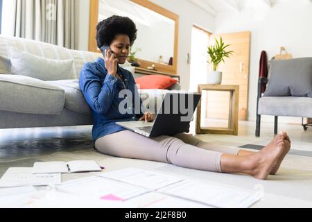 Mid adult african american female freelancer using laptop and talking on mobile phone at home Stock Photo