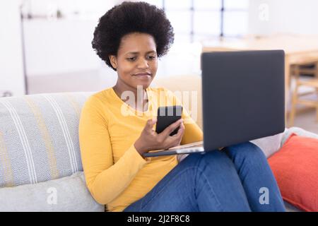 Smiling mid adult african american female freelancer using smart phone and laptop on sofa Stock Photo