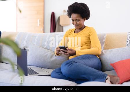 Smiling mid adult african american female freelancer using phone while sitting with laptop on sofa Stock Photo