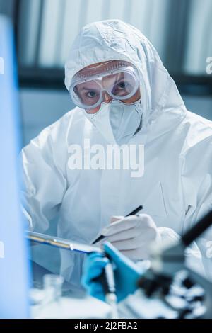 virologist in hazmat suit and goggles looking at camera on blurred foreground,stock image Stock Photo