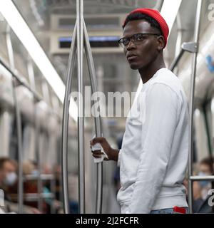 Portrait of thoughtful Black student man in red hat holding handrail with napkin in subway train. Stock Photo