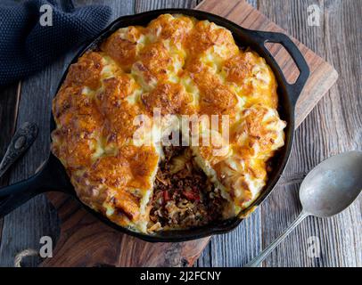 Savory pie with minced meat and cabbage. Topped with baked mash potato crust and cheddar cheese. Served in cast iron skillet on wooden table. Top view Stock Photo