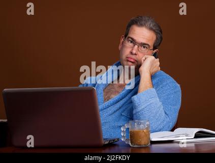 Middle aged man on the phone working at home in from of a laptop wearing a bathrobe and drinking a cup of coffee Stock Photo