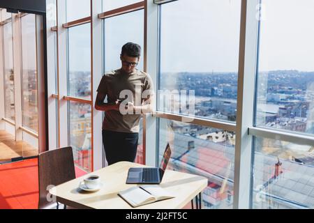 Cheerful scofe male entrepreneur standing near office window and smiling during working day in company, successful corporate boss feeling good from ri Stock Photo