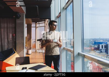 European man drinks coffee and looks out the window in a cafe. A young smiling student in glasses stands near the window. Recreation, leisure and free Stock Photo