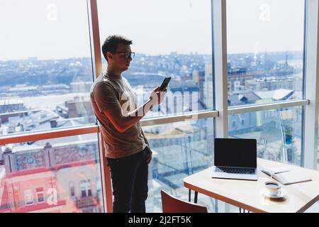 Cheerful scofe male entrepreneur standing near office window and smiling during working day in company, successful corporate boss feeling good from ri Stock Photo