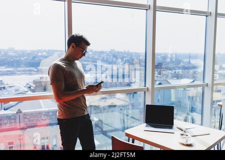 Cheerful scofe male entrepreneur standing near office window and smiling during working day in company, successful corporate boss feeling good from ri Stock Photo