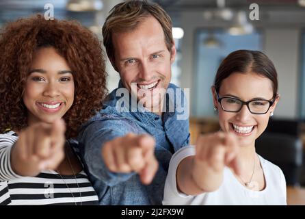 Were confident about you. Three friendly people pointing at you with their office space behind them. Stock Photo