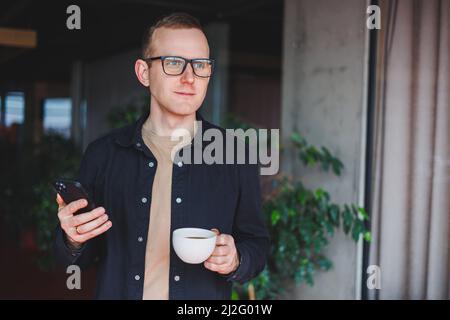 Portrait of a happy successful man in optical glasses holding a modern mobile phone and coffee in his hands, a cheerful blogger with a digital smartph Stock Photo