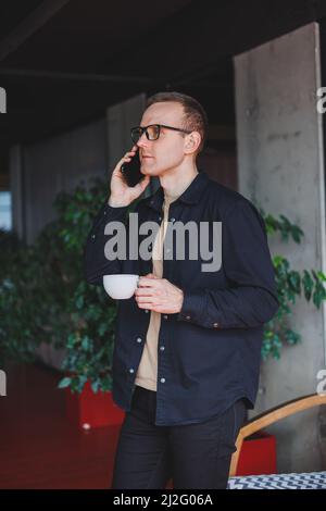 Portrait of a happy successful man in optical glasses holding a modern mobile phone and coffee in his hands, a cheerful blogger with a digital smartph Stock Photo