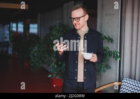 Portrait of a happy successful man in optical glasses holding a modern mobile phone and coffee in his hands, a cheerful blogger with a digital smartph Stock Photo