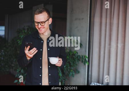 Portrait of a happy successful man in optical glasses holding a modern mobile phone and coffee in his hands, a cheerful blogger with a digital smartph Stock Photo
