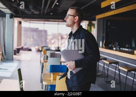 Male entrepreneur in black shirt and glasses standing in office with laptop in hand, successful corporate boss feeling good from rich lifestyle Stock Photo