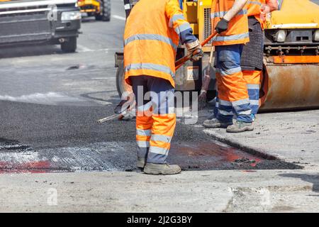 A group of road workers in orange overalls are repairing a section of the carriageway by rolling up fresh asphalt. Stock Photo