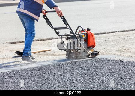 A road service worker uses a vibrating plate to compact asphalt at a road repair site on a sunny day. Copy space. Stock Photo