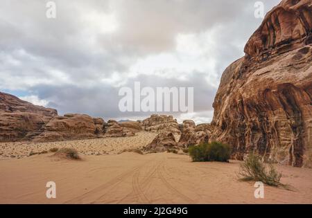 Rocky massifs on red orange sand desert, overcast sky in background, 4wd vehicle track print over sand - typical scenery in Wadi Rum, Jordan Stock Photo