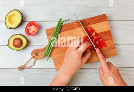 Avocado halves, spring onions, hand slicing tomatoes - basic guacamole ingredients on working board, view from above Stock Photo