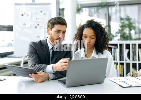 Successful colleagues, office workers, financial consultants, economists, sit in front of a laptop screen, in a modern office, develop a working strategy, analyze data, discuss the work concept Stock Photo