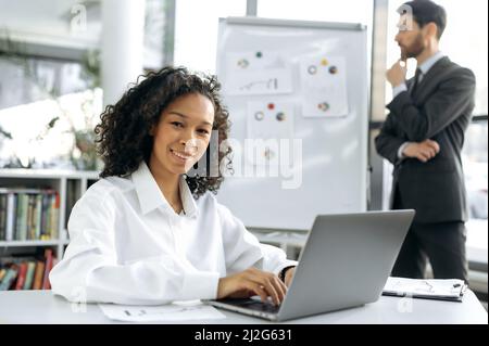 Successful African American woman sits at her desk in front of a laptop, looks at the camera, smiles. In the background, a male colleague works with charts, develops a strategy for modernizing work Stock Photo