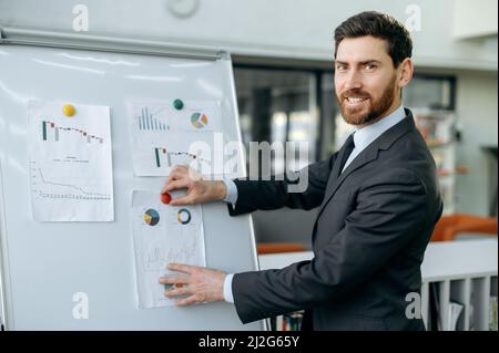 Handsome intelligent caucasian businessman, entrepreneur, stands near whiteboard with graphs, in modern office, presents financial forecasts and strategy of a new project, looks at camera, smiling Stock Photo