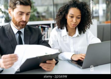 Concentrated successful business colleagues, a caucasian man and african american woman, co-founders of a company, work at a laptop, look through documents, discuss the company's development strategy Stock Photo