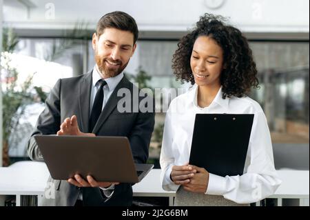 Positive friendly coworkers, a dark-skinned pretty woman and influential caucasian man, are standing in a modern office, viewing a report on a laptop, develop business plan and strategy, smiling Stock Photo
