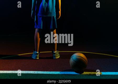 Basketball. A teenage boy in a blue sportswear stands on the Playground with ball. Close up of legs. Black background. Rear view. Concept of sports ga Stock Photo