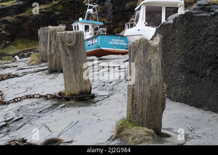 Rusty chain around the base of one of the wooden stakes used as  bollards on the quay in the harbour in Boscastle with a fishing boat hauled up in the Stock Photo