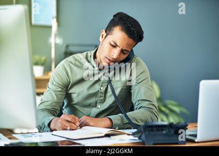 He is constantly busy dealing with clients. Shot of a focused young businessman seated at his desk while taking on the phone and making notes inside Stock Photo