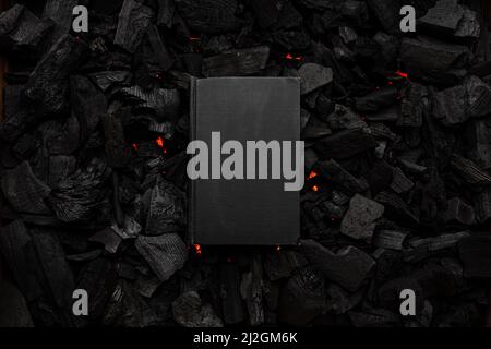 A closed book with a black cover on smoldering charcoal. Textured dark background. Stock Photo