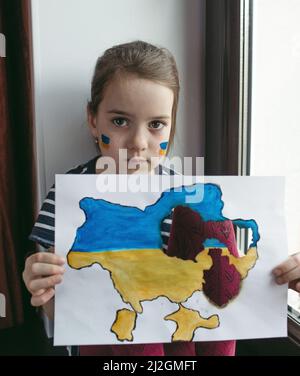 a little Ukrainian girl with tears in her eyes is sitting at the window holding a burnt map of Ukraine in her hands. Children and war Stock Photo