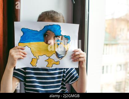 a little Ukrainian girl with tears in her eyes is sitting at the window holding a burnt map of Ukraine in her hands. Children and war Stock Photo