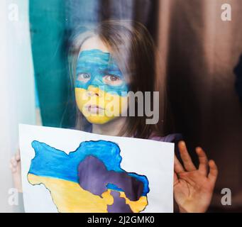 a little girl with a painted Ukrainian flag on her face is standing behind the glass with sad eyes holding a burnt map of Ukraine in her hands. Childr Stock Photo