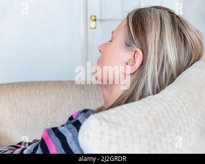 Female taking a break from work,listening to a language learning app,relaxing on the sofa with her eyes closed,in her living room,connected wirelessly Stock Photo