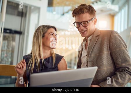 A young business woman is glad showing a laptop content to her young male colleague while standing in a pleasant atmosphere in the hallway together. B Stock Photo
