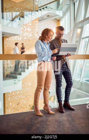 A business woman is watching a laptop content shown to her by young male colleague while standing in a pleasant atmosphere in company building hallway Stock Photo