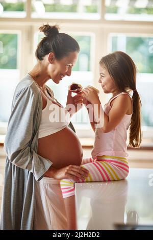 Sharing some sweet treats together. Shot of a pregnant woman and young girl eating cupcakes together. Stock Photo