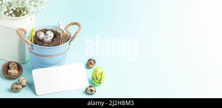 Nest of quail eggs in small bucket, small white gypsophila flowers, candle, empty white note on pastel blue backdrop. Festive Easter greeting card. Wi Stock Photo