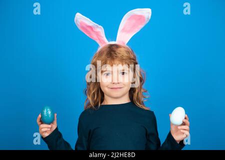 Child boy hunting easter eggs. Portrait of cute kid in rabbit costume with bunny ears having easter isolated on blue background. Funny kids face. Stock Photo