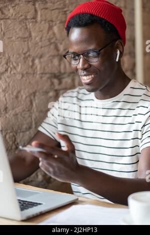 Smiling African freelancer man talking in video chat with headphones, remotely online work in cafe Stock Photo
