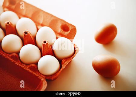 On a white table there is a red cardboard package from the store with white chicken eggs, and two brown chicken eggs are lying next to it. Healthy foo Stock Photo