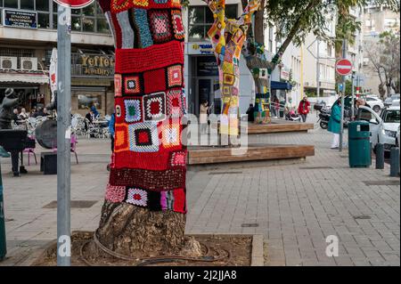 Netanya, Israel - February 7, 2022: Colorful crochet knit on a tree trunk yarn bombing. Patchwork knitted crochet covered tree for warmth, protection Stock Photo