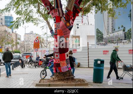 Netanya, Israel - February 7, 2022: Colorful crochet knit on a tree trunk yarn bombing. Patchwork knitted crochet covered tree for warmth, protection Stock Photo