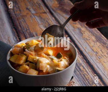 A white bowl of zucchini cubes with tomato soup, onions, garlic, condiments and melted cheese, served with a spoon over a rustic wooden table. Stock Photo