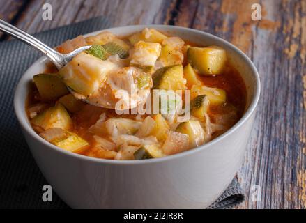 A white bowl of zucchini cubes with tomato soup, onions, garlic, condiments and melted cheese, served with a spoon over a rustic wooden table. Stock Photo