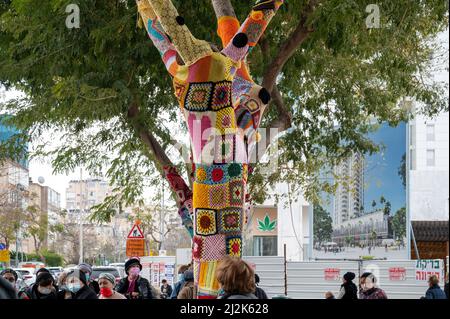 Netanya, Israel - February 7, 2022: Colorful crochet knit on a tree trunk yarn bombing. Patchwork knitted crochet covered tree for warmth, protection Stock Photo