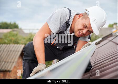 Man installer mounting photovoltaic solar moduls on roof of house. Close up electrician in helmet looking on evenness of solar panel during installing. Concept of alternative and renewable energy. Stock Photo