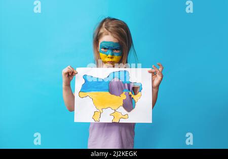 Portrait of a little girl with a painted Ukrainian flag on her face, standing on a blue background and holding a burnt map of Ukraine in her hands. Ch Stock Photo
