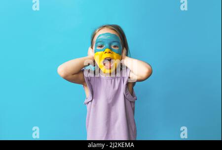 a little frightened girl with a Ukrainian flag painted on her face covers her ears with her hands and screams loudly against an isolated blue Stock Photo